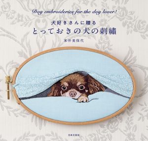 Cover of a Japanese dog embroidery patterns book showing a realistic silk shading embroidery of a Chihuahua peeking from under a blanket displayed in an oval wooden hoop.