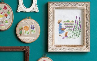 Seasonal cross stitch patterns book cover with finished hoops and framed pieces on a teal wall, including a lavender garden scene and floral swags