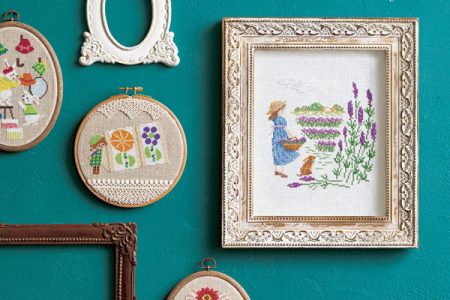 Seasonal Cross Stitch Patterns — 30+ Captivating Designs for Every Gift & Occasion Cover - Kayliebooks Seasonal cross stitch patterns book cover with finished hoops and framed pieces on a teal wall, including a lavender garden scene and floral swags