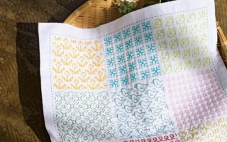Cover of a Japanese sashiko book showing a nine-pattern sashiko sampler cloth beside daisies in a woven tray