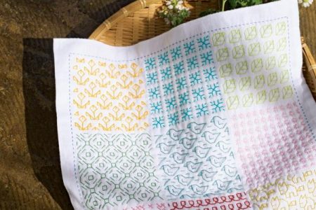 Cover of a Japanese sashiko book showing a nine-pattern sashiko sampler cloth beside daisies in a woven tray