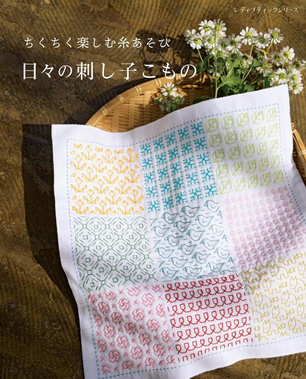Cover of a Japanese sashiko book showing a nine-pattern sashiko sampler cloth beside daisies in a woven tray